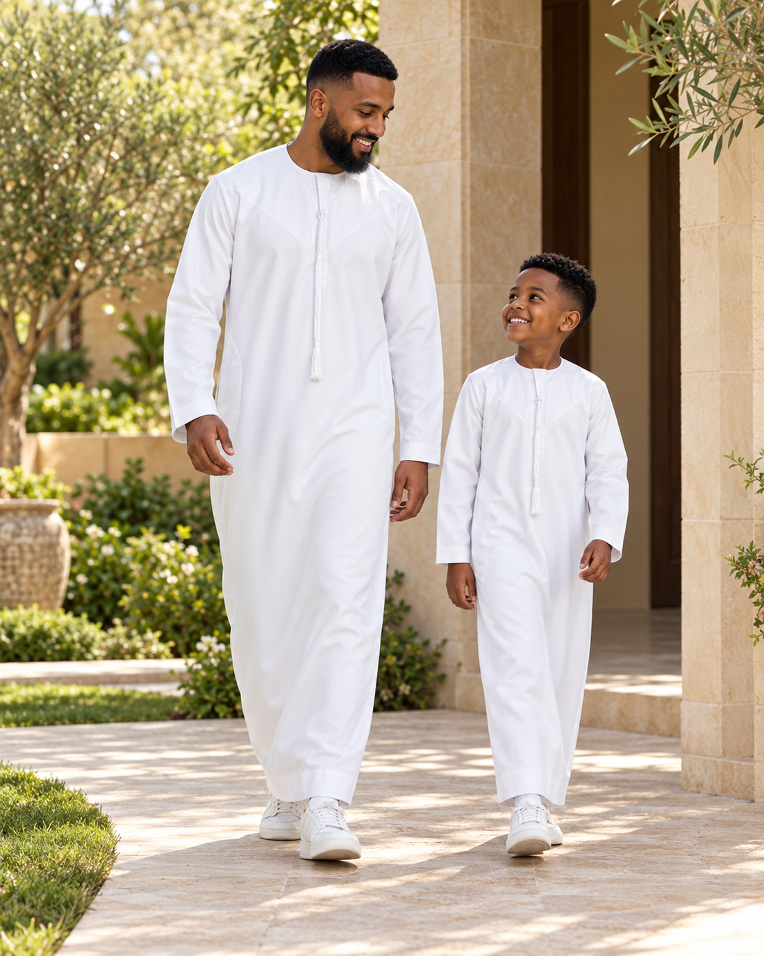A father and son wearing white tassel thobes and trainers, smiling at one another beside a garden doorway.